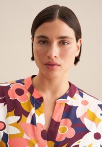Blumenbluse mit großen rosa und gelben Blumen auf lila Hintergrund, kurzen Ärmeln und einem Notch-Kragen mit einem pfirsichfarbenen Besatz.