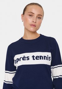 Mörkblå stickad tröja med texten "après tennis" i vitt över bröstet; vita ränder på ärmarna; rund halsringning; mjukt material.