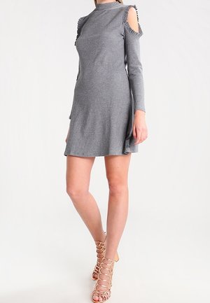 Vrouw draagt een grijze, lange mouwen, cold-shoulder gebreide jurk met ruchtrand en beige veterhoge sandalen, staand tegen een witte achtergrond.