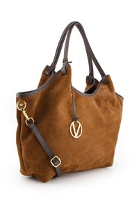 Bruine suède tote bag met donkere leren accenten, dubbele handvatten en een verstelbare schouderriem. Beschikt over een gouden logo charm en een zachte textuur.