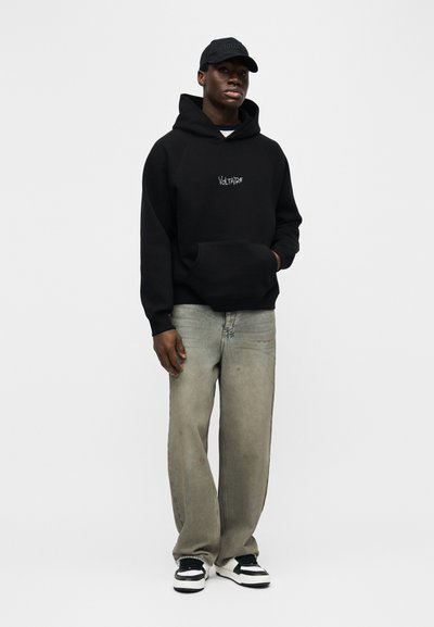 Άνδρας που φοράει μαύρο φούτερ με κουκούλα με το κείμενο "Voltaire", μαύρο καπέλο, φαρδιά ξεθωριασμένα γκρι τζιν και ασπρόμαυρα αθλητικά παπούτσια, στέκεται μπροστά σε απλό φόντο.