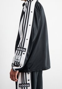 Svart sportjacka med vit tre-stripesdesign längs ärmarna, tryckknappar och en Adidas-logopatch på ärmen. Slät textur.