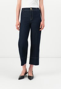 Žena v tmavě modrých širokých nohavicích ke kotníkům s dvojitým zapínáním a černých slingback špičatých podpatcích.