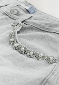 Szürke farmernadrág dekoratív díszítéssel, áttetsző és fehér gyöngyszerű díszekkel, amelyek texturált megjelenést kölcsönöznek a zsebeknek.