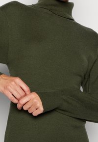 Pull col roulé vert olive avec une texture côtelée, manches longues et coupe ajustée. Gros plan sur des mains ajustant le tissu.