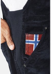 Marineblaue Corduroy-Latzhose mit einer auffälligen roten Tasche im blau-weißen Flaggen-Design, ergänzt durch strukturierten Stoff und kontrastierende Nähte.