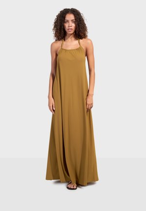 Vrouw met krullend haar, gekleed in een mosterdgele halternek maxijurk en zwarte sandalen, staand tegen een effen lichte achtergrond.