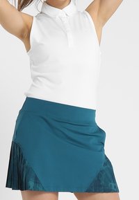 Bílá bezrukávová polokošile s límečkem, doplněná o tealovou sukni s nabíranými záhyby a potiskem na lemu.