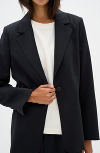 Black tailored blazer, structured shape, broad lapels, single button closure, textured fabric, worn over a white top.