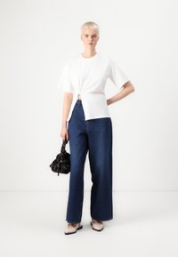 Tricou alb cu mâneci scurte și detaliu răsucit în față, asortat cu blugi largi de denim de culoare albastră închisă și pantofi plates de culoare deschisă. Geantă neagră.