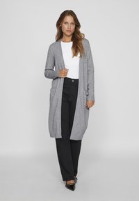 Long cardigan gris en tricot doux, avec un devant ouvert, des manches à bord côtes et deux poches avant, associé à un t-shirt blanc et un pantalon noir.