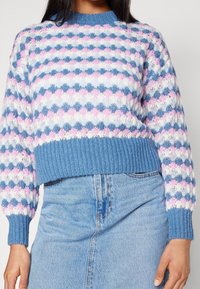 Persona vestida con un suéter de punto con un patrón de conchas en azul, rosa y blanco, y una falda de mezclilla azul clara, visible la sección media del torso.