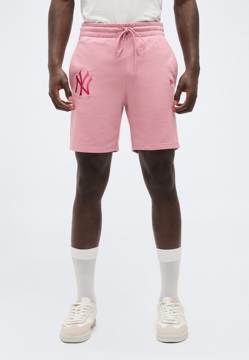 Mann trägt pinke Shorts mit einem pinkfarbenen New York Yankees-Logo, weiße Socken und weiße Turnschuhe, steht vor einem schlichten Hintergrund.