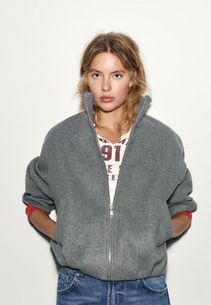 Junge Frau mit blonden Haaren trägt eine graue Reißverschlussjacke über einem weißen Shirt mit rotem Aufdruck und blauen Jeans, Hände in den Jackentaschen.