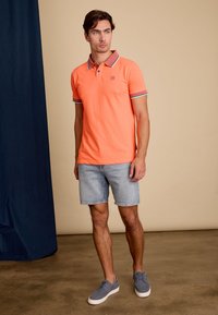 Oranje poloshirt met een contrasterende kraag, gedragen met lichtblauwe spijkerbroekshorts en grijze sneakers. Effen kleur, korte mouwen, casual pasvorm.