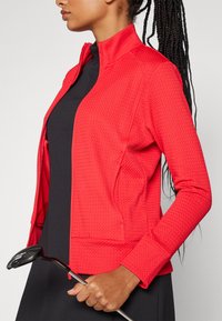 Casaquetão texturizado vermelho com gola, mangas longas e fecho de zíper. Usado sobre uma blusa preta, mão segurando um taco de golfe. Design limpo, estilo ajustado.