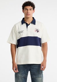 Polo shirt in beige with navy horizontal stripe, featuring a collar, short sleeves, and embroidered logo detail on the chest.
