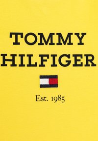 Żółta bawełniana koszulka z wyraźnym czarnym napisem "TOMMY HILFIGER" oraz małym haftowanym logo w kolorach czerwonym, białym i niebieskim. Napis zawiera "Est. 1985."