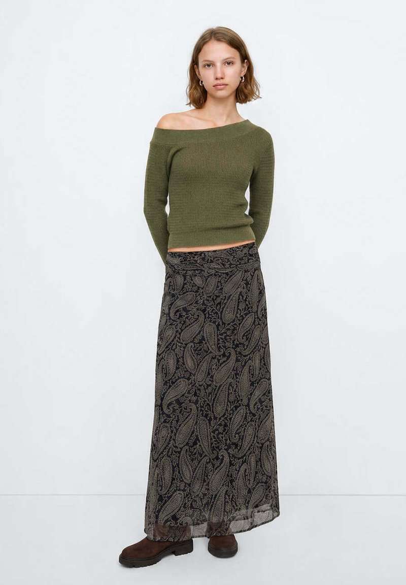 Jeune femme debout portant un pull vert olive épaules découvertes, une longue jupe noire à motif paisley et des bottes marron foncé sur un fond uni.