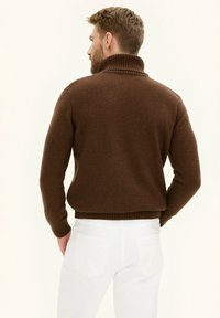 Maglione lavorato a maglia marrone con colletto alto a coste e fondo a coste, abbinato a jeans bianchi. Focalizzazione sulla texture e sul contrasto di colore.
