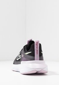 Bottine de sport en maille noire avec des accents violets et une semelle blanche. Comprend une languette au talon avec un logo et des détails réfléchissants sur le dessus.