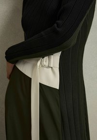 Zwarte geribbelde lange mouw top met een gestructureerde groene rok. Kenmerkt een witte riem met een ronde gesp en een knoopdetail.
