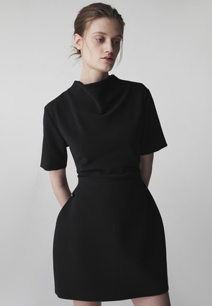 Jeune femme aux cheveux attachés, portant une robe noire à manches courtes avec un col bénitier, mains dans les poches, regardant vers le bas et de côté.