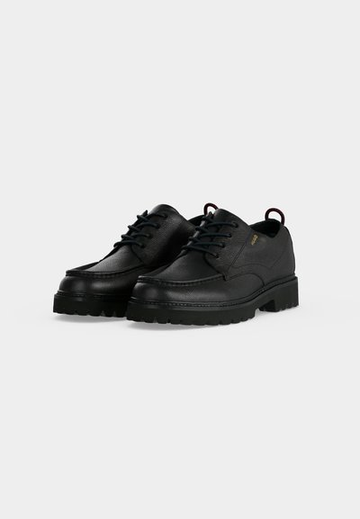 Chaussures en cuir noir à lacets avec surface texturée, semelles épaisses et robustes, et languettes aux talons sur un fond uni gris clair.