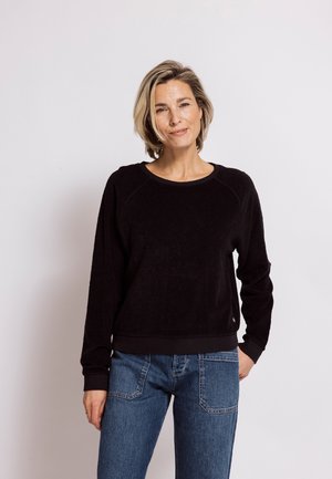 Woman with short blonde hair wearing a black long-sleeve shirt and blue jeans, standing against a plain light background.
