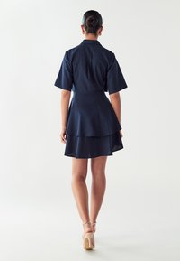 Tamsiai mėlyna sluoksniuota suknelė su trumpomis rankovėmis, prigludusia liemeniu ir išplatintu apvadu. Lygi tekstūra, sukurta laisvam prigludimui. Šviesus, šviesus fonas.