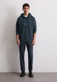 Sweat à capuche vert foncé avec un motif sur le devant, des cordons de serrage et des poignets côtelés, associé à un pantalon noir et des chaussures noires, se tenant devant un fond neutre.
