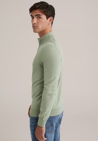 Világoszöld pulóver, magas gallérral, testhezálló kialakítással és bordázott mandzsettákkal. Puha, sima anyagból készült, minimális textúrával.