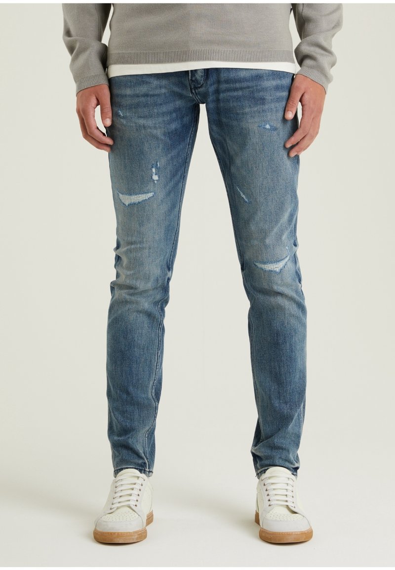 Jeans de mezclilla azul claro con detalles desgastados, con un ajuste ceñido, lavado desvaído y un ligero desgarro en las rodillas, combinados con zapatillas blancas.