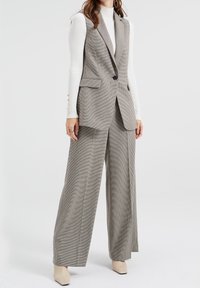 Šedá vesta s kostkovaným vzorem a dvěma předními kapsami, doplněná o široké kalhoty se stejným vzorem. K outfitu patří bílý žebrovaný top s dlouhým rukávem pod vestou.