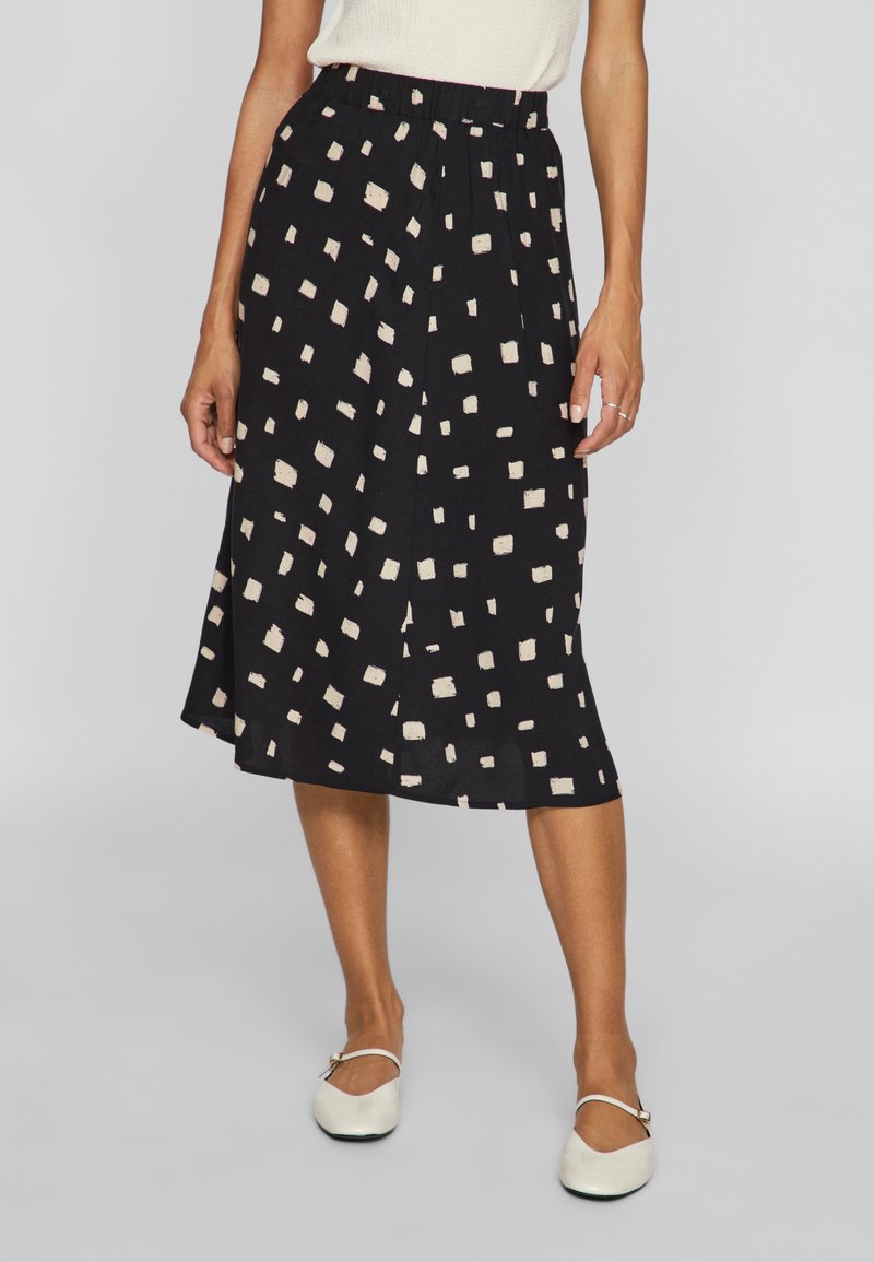 Femme portant une jupe midi noire avec des motifs carrés beiges, un haut blanc, des chaussures Mary Jane blanches, debout devant un fond gris uni.