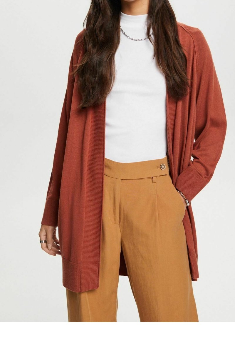 Cardigan couleur rouille en maille côtelée, design ouvert à l'avant ; associé à un haut blanc et un pantalon taille haute marron avec un détail bouton.