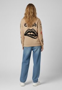 Felpa beige con un grafico di labbra nere e testo sulla schiena, abbinata a jeans larghi azzurri chiari e sneakers bianche.