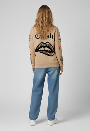 Felpa beige con un grafico di labbra nere e testo sulla schiena, abbinata a jeans larghi azzurri chiari e sneakers bianche.