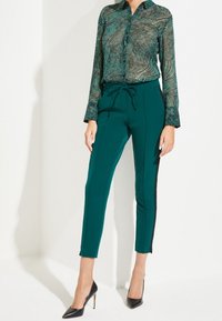 Groene blouse met een patroon en een kraaghalslijn, gecombineerd met teal tapered broek met zijkanten in zwart en zwarte hakken.