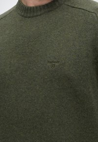 Gros plan sur une personne portant un pull en maille vert foncé avec un col rond côtelé et un petit logo brodé sur la poitrine.