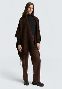 Brown leopard print poncho with fringed edges, made of soft fabric. Worn over a black top and paired with brown trousers and black boots.