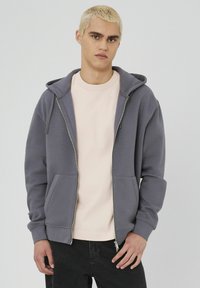 Un sweat à capuche gris zippé avec une grande capuche, des poches avant et des poignets côtelés ; porté sur un t-shirt rose pâle, présenté sur un fond neutre.