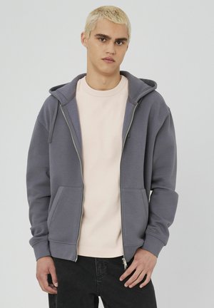 Un sweat à capuche gris zippé avec une grande capuche, des poches avant et des poignets côtelés ; porté sur un t-shirt rose pâle, présenté sur un fond neutre.