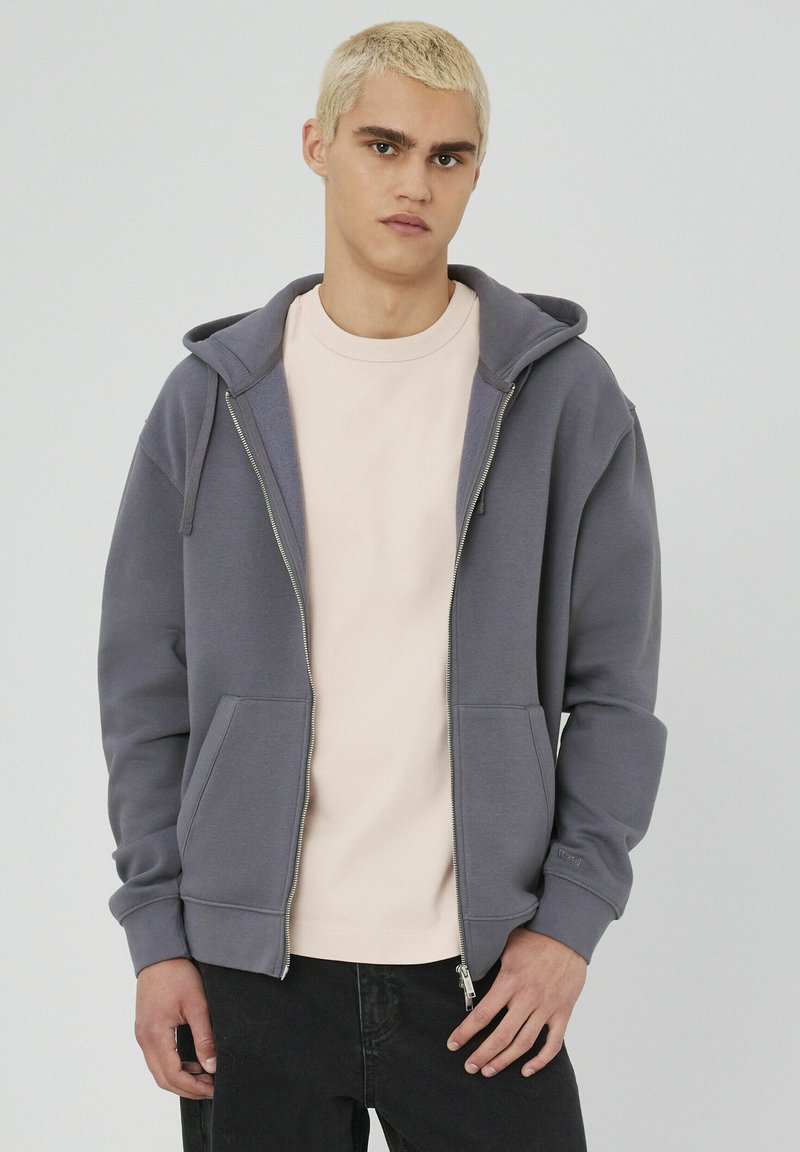 Un sweat à capuche gris zippé avec une grande capuche, des poches avant et des poignets côtelés ; porté sur un t-shirt rose pâle, présenté sur un fond neutre.