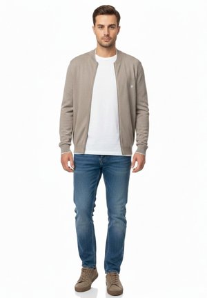 Hombre de pie mirando hacia adelante, vestido con chaqueta beige con cremallera, camiseta blanca, vaqueros azules y zapatillas beige sobre fondo blanco.