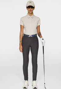 Donna con polo color beige chiaro e cappellino, pantaloni scuri, guanti bianchi e sneakers, che tiene un bastone da golf davanti a uno sfondo neutro.