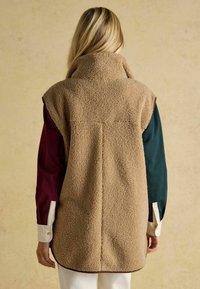 Beige Fleece-Weste mit hohem Kragen, ärmellosem Design und strukturierter Oberfläche. Kanten sind braun gesäumt, darunter ist ein mehrfarbiges Hemd zu sehen.