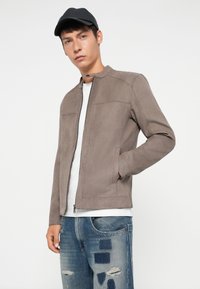 Chaqueta de ante caqui con cuello alto, cierre frontal de cremallera, dos bolsillos laterales y una textura suave. Usada sobre una camiseta blanca y jeans desgastados.