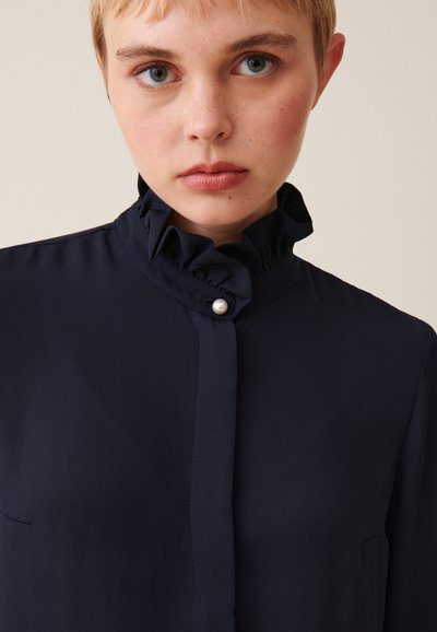Blusa blu navy con colletto a volant, chiusura con bottoni perla e una texture liscia con un leggero brillantezza. Maniche lunghe con dettagli minimi.