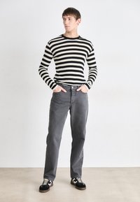 Chemise à rayures noires et blanches à manches longues, pantalon gris et baskets noires avec accents blancs, se tenant sur un fond neutre.